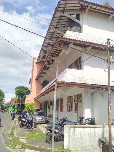 The image shows a two-story building with a white exterior and a red roof located on a street in Kecamatan Kartasura, Indonesia (latitude: -7.565, longitude: 110.776). The building appears to be a commercial or institutional structure. The building has windows and a balcony on the second level. There are multiple motorbikes parked in front of the building, and two people can be seen standing near the motorbikes. One person is wearing a blue shirt, and the other person is wearing a green shirt. In the background, a car is parked across the street. The street is paved and has a sidewalk on both sides. A single, small tree is visible in the foreground, while several larger trees are visible in the background. Power lines run overhead throughout the scene. The sky is mostly clear, with a few white clouds.