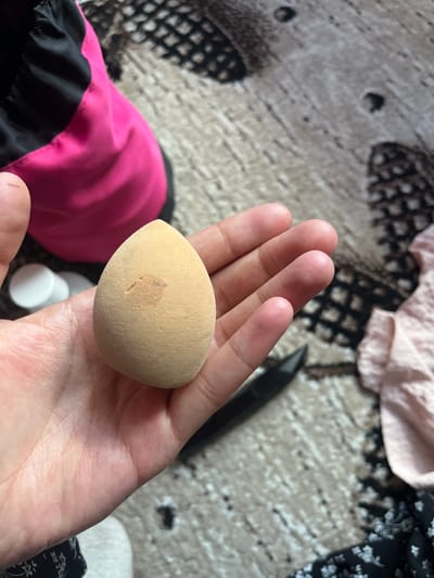 A hand holds a light brown, egg-shaped makeup sponge. The sponge has a slight discoloration, appearing as a darker spot near the center. The hand is fair-skinned and appears to be that of an adult. In the background, a pink and black garment is visible, likely part of a jacket or bag. Several small white circular objects, possibly small containers or lids, are also present in the lower right portion of the frame. The surface beneath the objects is a textured rug or carpet with a pattern of dark and light grey shapes. A sliver of black object, possibly a makeup brush or pencil, is visible in the upper right background. The lighting suggests it is daytime, with natural light illuminating the scene. The overall impression is of a casual setting, perhaps a room in a home. There is no visible text in the image.