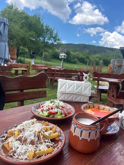 The image depicts an outdoor dining scene, likely in a restaurant or picnic area, located in Meshtitsa, Bulgaria. A wooden table is set with traditional Bulgarian dishes, including a plate of French fries topped with shredded cheese, a plate of sliced tomatoes and cucumbers, and a ceramic pitcher filled with a white liquid. A white quilted handbag with a decorative handle rests on the table. In the background, wooden tables and benches suggest an outdoor seating arrangement, and a lush green landscape with trees and a distant mountain under a partly cloudy sky can be seen. The style of the dishes and the surrounding environment suggest a focus on local cuisine and natural scenery.