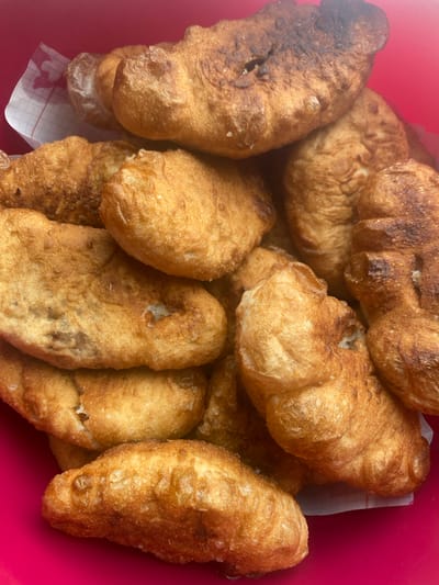 A close-up, overhead shot shows a pile of fried dough pastries, likely belyashi, in a bright pink bowl. The pastries are irregular in shape and golden brown with some darker, more caramelized spots. They appear to be soft and slightly greasy. A piece of white checkered paper is visible under some of the pastries on the left side of the frame. The background is a solid, vibrant pink. The image does not contain any information about the city of Бетюнцы, Russia, or any people or actions.