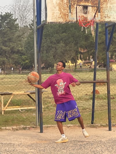 An outdoor basketball court in Umunekwu, Nigeria, features a single male subject, appearing late teens to early twenties, with dark skin and short dark hair. He wears a magenta t-shirt displaying a reddish-brown robot graphic, purple basketball shorts with "LAKERS" in gold lettering, and light-colored athletic footwear. He holds a worn, orange basketball in his left hand, with his arm extended. His head is tilted back, eyes squinted or closed, and mouth closed, suggesting concentration. He is captured in mid-action, balancing on his left foot with his right foot slightly lifted. Behind the subject, a heavily rusted basketball backboard with a red rim and a white, frayed net is visible, supported by blue poles. A chain-link fence defines the court boundary, behind which are a grassy field, numerous green trees, some bare deciduous trees, and faint human figures. A light-colored wooden bench-like structure is present to the left. The court surface is worn, light-colored asphalt or concrete.