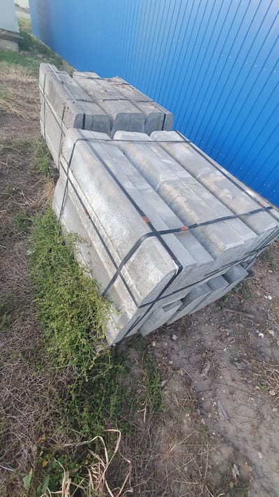 The image depicts a stack of concrete curbs placed outdoors, with a blue corrugated metal wall in the background. The curbs are gray, rectangular with a curved top edge, and are banded together with black plastic straps. They are stacked in two main piles, with one pile partially obscured by green vegetation. The ground around the curbs is covered with dry grass, dirt, and small stones. The overall scene suggests an outdoor storage area, possibly at a construction site or material yard.