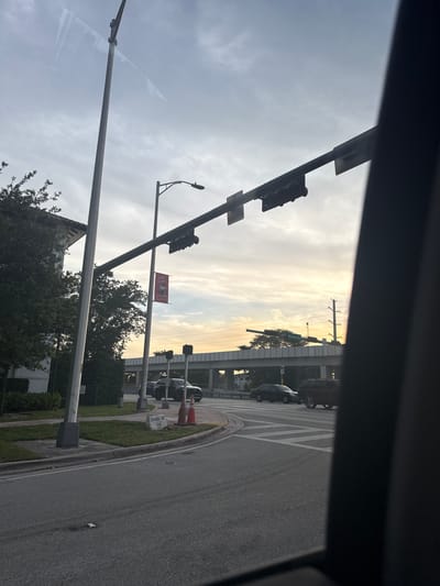 The image depicts a street scene in Coral Gables, Florida, USA, during what appears to be sunset or sunrise. In the foreground, a street with crosswalk markings is visible, with a curb and a grassy median to the left. A traffic light pole stands prominently on the left side of the image, and another traffic light is suspended over the intersection. A banner that reads "South Gables" with floral imagery is hanging from a pole. Several cars are present in the intersection and on the road. In the background, an elevated highway or overpass is visible. The sky is cloudy with a warm glow indicating low sunlight. The overall scene suggests an urban environment with traffic and infrastructure.