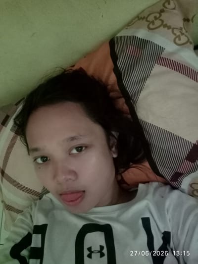 A young female lies on her back, propped up by pillows. She is wearing a white t-shirt with a black logo and has her tongue sticking out, facing the camera. The background appears to be a wall in a bedroom setting, with a patterned pillow visible to her right. The timestamp on the image indicates it was taken on June 27, 2026, at 13:15. The location is identified as Sukaraja, Indonesia.