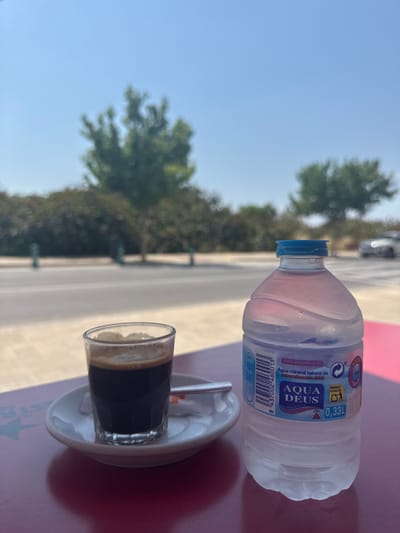 The image shows a glass of dark liquid, likely coffee, on a white saucer with a small metal spoon, and a partially consumed bottle of "Aqua Deus" mineral water, on a table with a pink-red surface. The water bottle's label is partially visible, showing the brand name, volume (0.33L), and recycling information. The background features a blurred street scene with trees and a bright blue sky, suggesting an outdoor setting. Given the brand of water and the overall environment, the location is very likely in Vinaròs, Spain, given that it is a Spanish brand of water. The perspective suggests the photo was taken from a seated position at the table.
