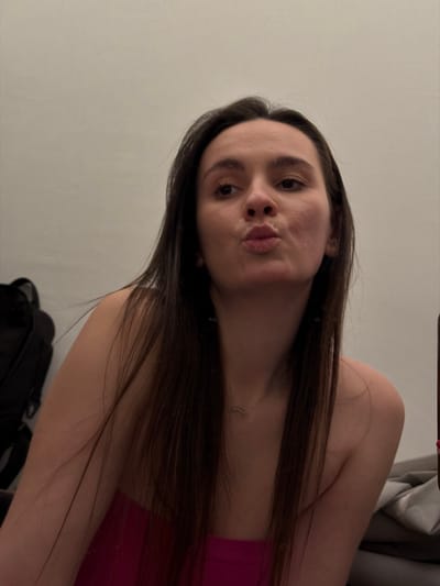 A young woman with long, dark brown hair is posing with puckered lips. She is wearing a hot pink strapless top. A delicate necklace with a small rectangular pendant is visible around her neck. The background includes a light gray wall and the corner of what appears to be a gray upholstered couch or chair. A black bag is positioned in the lower part of the frame.