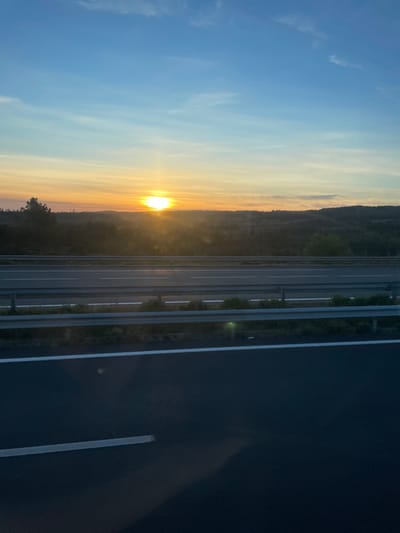 The image depicts a multi-lane highway scene near Sigean, France, during either sunrise or sunset. A vibrant, spherical sun is positioned low on the horizon, slightly left of center, emitting strong golden light that casts a warm glow across the landscape and reflects faintly on the asphalt. The sky above transitions from clear blue to warm orange and yellow hues near the horizon, featuring scattered cirrus clouds. The background consists of gently rolling hills densely covered in dark green foliage, with a slender, vertical structure silhouetted on the far left horizon. In the midground, a dark asphalt highway features multiple lanes delineated by crisp white painted markings. A two-tier gray metal guardrail separates the immediate highway lanes from a vegetated median or roadside. No individuals, vehicles, or specific activities are discernible within the frame.