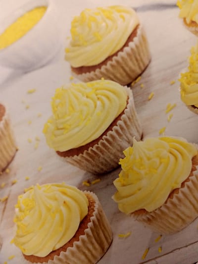 The image displays a collection of yellow-frosted cupcakes with yellow sprinkles, arranged on a light-colored surface that appears to be wooden planks. In the background, a white bowl filled with a yellow substance, possibly frosting or a sauce, is partially visible. The lighting is soft, creating a slightly blurred effect, particularly in the background. There are no people present in the image, and no discernable activity is depicted. The scene is a close-up of desserts, implying a food-related context. There is no information within the image that indicates the location of Rumaithiya, Kuwait.