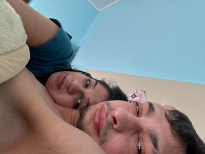 The image displays two individuals, an adult male and an adult female, lying down in an indoor setting within the city of Касапите, Bulgaria. The male is in the foreground, his face and upper torso visible, featuring dark hair, a mustache, and a short beard. His eyes are open and directed towards the camera. The female is positioned behind him, with her head resting close to his arm or shoulder. She has dark hair, is wearing a dark teal-colored top, and her eyes are also open, looking towards the camera. A light-colored, soft object, possibly a pillow, is partially visible near the female's head and arm on the upper left. The room's environment includes a light blue wall in the upper and left sections, a beige wall on the right and bottom, and a white ceiling trim visible in the upper left corner. A patterned fabric, appearing pink with white and dark elements, is partially visible behind the male's head.