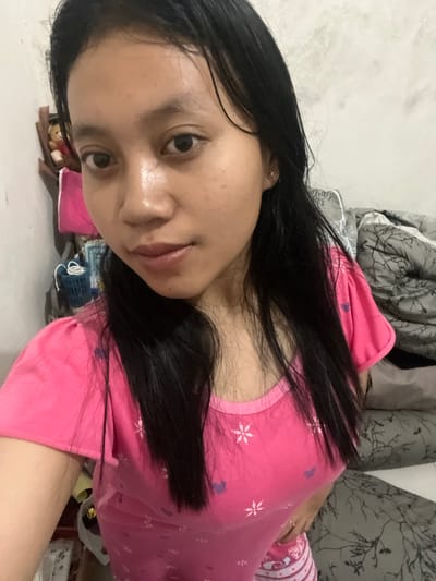 An eye-level, medium close-up selfie shows a young woman with straight, black hair framing her face and falling past her shoulders. She is wearing a bright pink t-shirt with a pattern of small white flowers and purple heart shapes. Her skin is moist, possibly from sweat or water. She has dark eyes and a neutral expression. The background is out of focus, but appears to be an indoor setting with a white wall on the left and a pile of gray and white patterned bedding on the right. A brown stuffed bear is partially visible on a shelf to the left of the woman. The scene is likely a bedroom. The provided location is Jambearum, Indonesia.