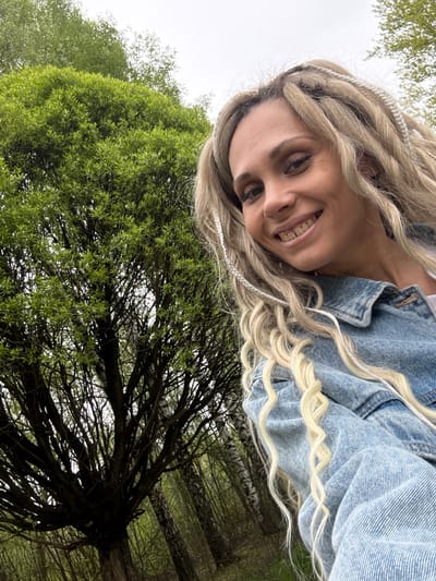 A woman with blonde dreadlocks and wearing a denim jacket smiles at the camera. The background includes green trees and a cloudy sky. This photo was likely taken outdoors in Dumino, Russia.