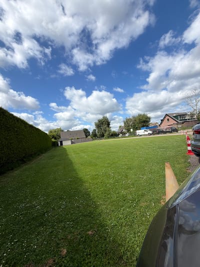 The image shows a grassy area with a large hedge on the left side. In the background, there are several houses and a street with parked cars and vans. The sky is blue with white clouds. The scene appears to be a residential area in Maarssen, Netherlands. There are no people visible in the image. The focus is on the outdoor environment.
