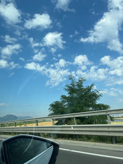 This media file captures a view from a vehicle, likely traveling on a highway near Brașov, Romania. The scene is dominated by a bright blue sky filled with fluffy white clouds. A faint contrail from an aircraft is visible stretching across the sky.
In the foreground, the side mirror of the vehicle is reflected, offering a glimpse of the surrounding landscape. A metal guardrail runs along the side of the road, separating it from a grassy verge and a dense green tree. Beyond the guardrail, a field of dry, golden grass stretches towards distant, rolling hills or mountains, shrouded in a light haze. A few structures, possibly buildings or industrial elements, can be seen faintly in the distance.
The time of day appears to be daytime, with bright sunlight indicating fair weather. The image evokes a sense of travel and open road, with the vast sky and distant landscape suggesting a journey through a rural or semi-rural area. There is no visible text in the media file.