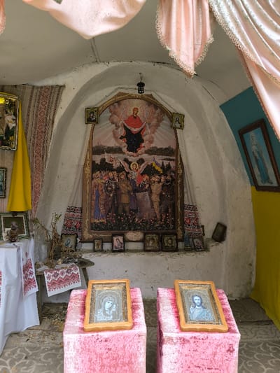 This image depicts the interior of a small, arched, white-washed structure, likely a chapel or shrine, located in Horaivka, Ukraine.
The central feature on the rear wall is a large, vertically oriented, gold-framed religious painting. The upper portion of this painting displays a figure in red and dark robes, possibly the Virgin Mary, suspended within clouds. The lower portion depicts a gathering of figures, including an angel with wings, individuals in military uniforms (some appearing contemporary), and others in civilian attire, standing in a field. Two Ukrainian flags are visible within this crowd. Flanking this primary painting, at a higher elevation, are two smaller, framed religious icons. Below the main painting, a narrow ledge holds a horizontal row of multiple small, framed icons. A long, white textile with red embroidered patterns hangs vertically behind the central painting. A dark, lantern-like fixture is suspended from the arched ceiling directly in front of the main painting.
To the left of the central arrangement, a wooden table or bench is partially covered by a white textile with red embroidered designs. On this surface, dried plant material and other small objects are present. Above and to the left of the table, another framed image is partially visible behind a draped yellow fabric.
The right wall is divided horizontally, with a light blue upper section and a yellow lower section. A framed image of the Virgin Mary, depicted in blue tones, hangs on the upper blue section. A small, dark framed object leans against the wall below.
In the foreground, two rectangular pedestals, covered in pink, textured fabric, stand centrally. Each supports a framed religious icon tilted towards the viewer. The icon on the left depicts the Virgin Mary holding the infant Jesus. The icon on the right depicts Jesus Christ. Pink, ruffled textiles also drape from the upper curved ceiling sections on both the left and right. The floor in the foreground displays a patterned surface.