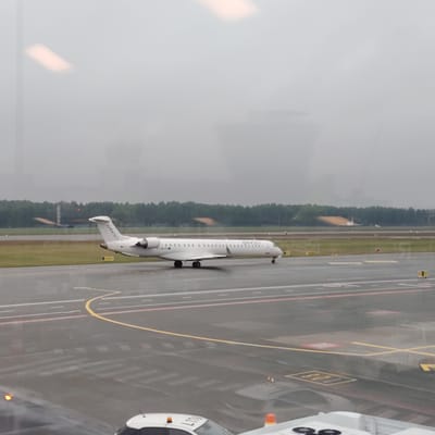 The image depicts an airport tarmac scene observed through a reflective window under overcast conditions, ostensibly located in Skulte, Latvia. A white CityJet regional jet aircraft, likely a Bombardier CRJ series, occupies the middle ground, positioned on a wet asphalt taxiway. The aircraft features "CITYJET" branding on its fuselage and vertical stabilizer, and a partial registration beginning with "EI-" is visible on the aft fuselage. It is equipped with two rear-mounted engines and a T-tail configuration.

The tarmac surface is visibly wet, reflecting ambient light and contributing to a subdued, grey illumination. Distinct yellow and white painted lines mark the taxiway boundaries and indications, with additional red markings present on the paved surface. Beyond the taxiway, green grass verges transition to a dense treeline that defines the distant horizon. In the far background, behind the trees, faint, blurry structures are discernible, including a lattice tower with red lights and another indistinct, light-colored object possibly an aircraft or airport infrastructure.

The immediate foreground and upper portions of the image exhibit significant reflections on the viewing window. A bright, rectangular indoor light is prominently reflected in the upper left. In the lower foreground, reflections include the upper portion of a white ground vehicle with an orange beacon, displaying the partial alphanumeric marking "L170" on its side, and another orange beacon to its right. A grey structural element, possibly part of the viewing area's interior, is also reflected in the bottom right corner. No individuals are clearly identifiable within the frame.