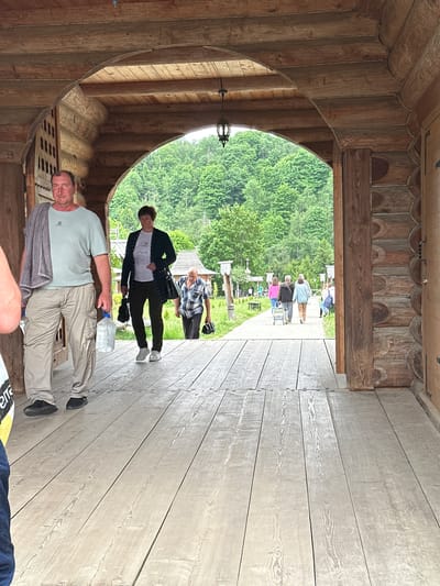 The image captures a scene in Vzglyadnevo, Russia, featuring a wooden structure with an arched doorway. Inside the structure, a man in a light green t-shirt and beige pants stands, holding a bottle. A gray jacket is draped over his shoulder. Beyond the doorway, several other people are visible in a grassy area with some trees. The building's interior shows detailed wooden construction with decorative elements. The floor appears to be made of light-colored wooden planks.