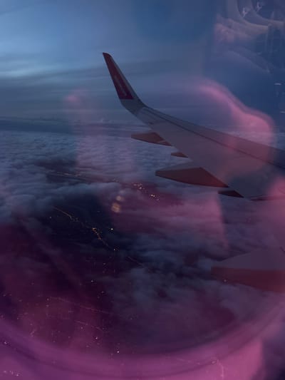 The image captures an aerial view from an airplane window, depicting the activity of flying over a landscape at night or during late twilight. A portion of an airplane wing is prominently visible, extending diagonally from the upper-left towards the middle of the frame. The wing's red-tipped end, located on the far left, clearly displays the text "wizzair.com", identifying the airline as Wizz Air.

Below the wing and extending across the right half of the image, dense layers of clouds partially obscure the ground. Through breaks in the clouds, numerous yellow and orange lights of what appears to be a city or populated area are visible, creating glowing patterns against the dark backdrop of the night sky.

A strong, translucent purple and pink wash overlays the entire scene, particularly intense on the right side, giving the image an abstract, ethereal quality. This distinctive color treatment blends with reflections, possibly from inside the aircraft cabin, visible as wavy forms in the upper-left. The blend of natural elements with the artistic purple haze creates a dreamlike perspective of the world from above, likely over a region such as Szentendre, Hungary, given the provided context.