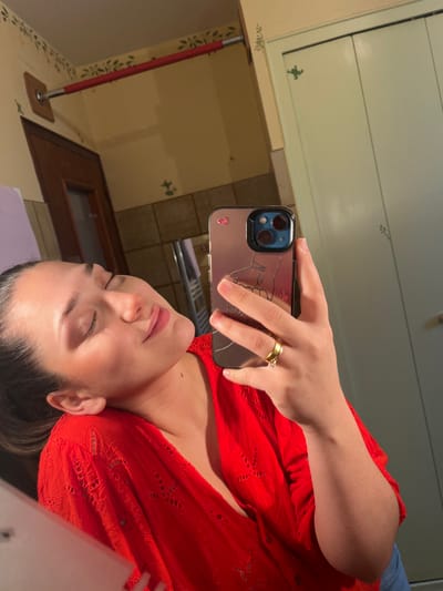 An image, identified as taken in Ancenis, France, depicts a mirror selfie of a female individual. The individual, positioned off-center to the left, has dark hair pulled back and wears a red, short-sleeved top with eyelet embroidery. Her eyes are closed, and her head is tilted slightly back and to the right, exhibiting a subtle smile or serene expression. She holds a smartphone in her right hand, reflected in the mirror. The phone case is metallic-toned, shows a prominent crack, and features a minimalist line art drawing of an upper body, along with small pink butterfly stickers near the camera module. A gold-colored band with a single clear stone is visible on her right ring finger. The background, also a reflection, reveals an interior space, likely a bathroom. The walls are light beige with a green stencil pattern depicting small floral or leaf motifs. A red horizontal bar is mounted on the upper left wall, secured by a wooden backplate. To the right, a light-colored, possibly green or white, two-panel door or cabinet with similar stencil details is visible. A metallic, ladder-style fixture, consistent with a towel warmer or radiator, is partially visible in the lower central reflected area.