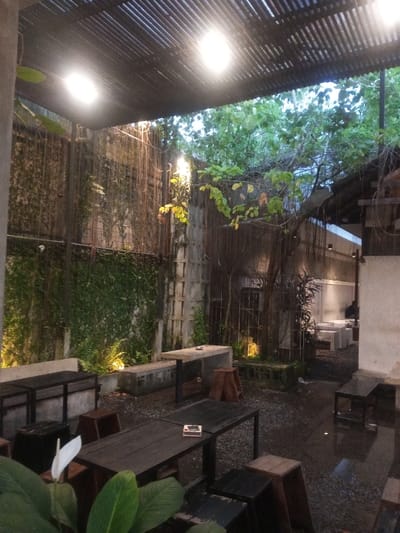 The image depicts a semi-outdoor seating area, characteristic of a cafe or restaurant, located in Pontianak, Indonesia. The scene is illuminated by artificial overhead lighting from two prominent square fixtures mounted under a dark, slatted roof, and by accent lights positioned at ground level along the walls and around plants. The time of day appears to be twilight or early evening, with ambient light visible in the sky above the roofline.
The ground is composed of dark, wet gravel or small pebbles. Multiple dark, rectangular tables and various brown wooden stools or low benches are arranged across the gravel. Integrated concrete benches are present along a tall, textured wall on the left, which is heavily covered in green climbing vines and foliage, with additional ferns in planters below. A large tree, possibly a banyan due to visible aerial roots, stands in the mid-ground, surrounded by concrete blocks and subtly lit from below.
In the background to the right, a lighter-colored building or wall section features additional seating arrangements, including white or light-toned blocks serving as tables and seats. A single, indistinct individual is faintly visible seated at one of these structures in the far background. The immediate foreground on the left displays a white flower, resembling a peace lily, with large green leaves. No direct interactions or specific actions are discernible beyond the general arrangement of a functional seating area.