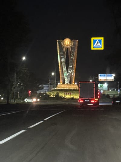 The image depicts a night scene in Leninavan, Russia, focusing on a monument at an intersection. A large, abstract metal sculpture stands prominently, illuminated by spotlights. To its right, a pedestrian crossing sign is visible. The foreground shows a paved road with white lane markings. A red-lit truck is driving away from the camera on the road, and a white car with its headlights on is seen in the distance, partially obscured by trees and streetlights. The overall environment is dark, characteristic of nighttime in an urban area, with various lights from street lamps, vehicles, and the monument illuminating the scene.