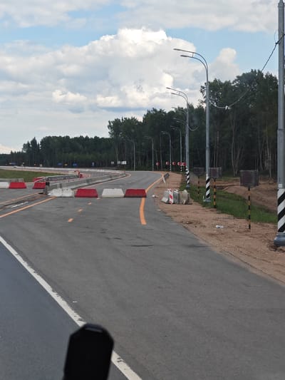 The image depicts a section of a road construction site in Khotilovo, Russia. The road itself is asphalt, with white and yellow lane markings. Several red and white concrete barriers are placed along the road, likely to redirect traffic or delineate construction zones. In the background, a dense line of trees forms the edge of the road. Several streetlights with multiple arms are visible, as are some traffic signs and poles with reflective black and yellow stripes. The sky above is partly cloudy with white cumulus clouds. The overall impression is that of ongoing infrastructure development.