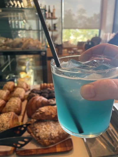 A clear glass containing a vibrant blue, ice-filled beverage with a dark straw is held by a human hand in the foreground. In the mid-ground, out of focus, a display of various baked goods including croissants and seed-topped pastries is visible on wooden boards. The background reveals an indoor commercial setting, likely a cafe or bakery in Nes Ziona, Israel, featuring a glass display case, shelves with bottles, and a large window providing natural light and an obscured view of outdoor foliage.
