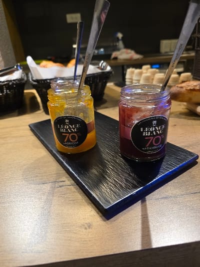 A close-up shot presents two small glass jars of jam or preserves, each with a spoon inserted. The top jar contains a dark red, raspberry-flavored preserve with a "Leonce Blanc 70%" label. The bottom jar holds a vibrant orange, apricot-flavored preserve, also labeled "Leonce Blanc 70%". Both jars sit on a textured, dark gray, possibly slate or metal, serving tray. The tray is angled on a wooden surface. The spoons read "pro mundi-INOX" on the handles.