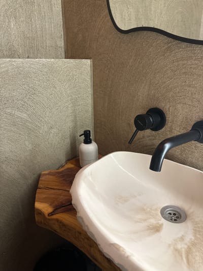 This image depicts a detailed view of a contemporary bathroom sink area, located in Усады, Russia. A prominent white ceramic vessel sink, displaying significant light brown and beige discoloration or staining within its basin, is the central object. A circular silver drain grate is visible at the bottom of the sink. The sink is supported by a live-edge wooden slab countertop, characterized by its natural brown tones, distinct wood grain, and visible cracks. Above the sink, a matte black wall-mounted faucet assembly consists of a curved spout and a single lever mixer handle, on which the text "H STIEWERKI C" is discernible. To the left of the sink, resting on the wooden countertop, is a light grey or beige cylindrical soap dispenser with a black pump. The surrounding walls are textured with a swirled or brushed pattern. The wall behind the faucet is light brown, while adjacent wall sections to the left feature a similar textured pattern in a lighter, greyish-beige hue, appearing as distinct panels. A segment of a mirror with a dark, wavy frame is visible in the upper right portion of the image, reflecting a portion of the textured wall.