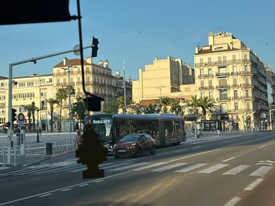 The image depicts a street scene in Toulon, France, likely on a sunny day given the clear blue sky and bright lighting. In the foreground, a paved road with white lane markings and a pedestrian crossing is visible. A dark silhouette of an air freshener, shaped like a pine tree, hangs from the viewpoint, obscuring a portion of the scene.

Dominating the mid-ground are a bus and a car. The bus, an articulated vehicle with "1 Beaucaire" displayed on its front, appears to be stopped at a bus stop. Next to the bus, a red Citroën C3 is positioned, facing forward. The license plate of the car is visible as "FD-218-DH".

The background features a street lined with multi-story buildings displaying classic European architecture with balconies and shuttered windows. Palm trees are scattered throughout the scene, adding a Mediterranean feel. A traffic light with a red light illuminated is visible on the left side of the image, along with a street sign. Several people are walking and standing on the sidewalk. A trash bin is also present on the sidewalk. The overall impression is of an urban environment in Southern France.