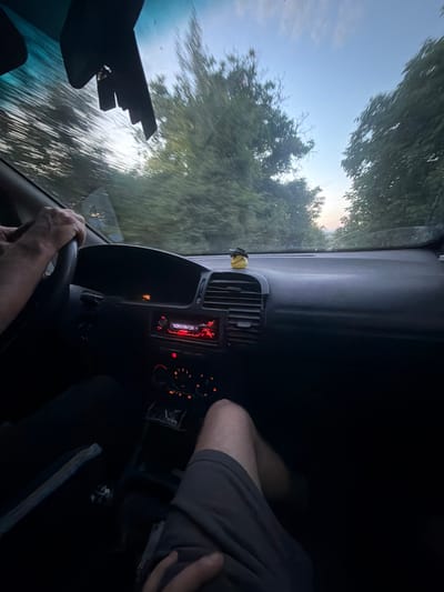 The image shows the interior of a car moving along a road lined with trees, likely taken from the driver's seat. The landscape is blurred due to the motion. The driver's hands are on the steering wheel. A passenger is sitting in the passenger seat. The car's dashboard and console are visible, including the radio and climate control panel. It is not possible to determine if this photo was taken in Kralevo, Bulgaria, or any other specific location.