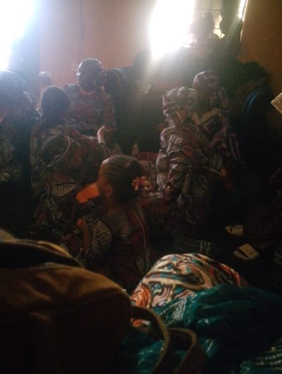 Several people, mostly women and one child, are seated in a brightly lit room. They wear colorful patterned clothing. Some wear headwraps. A tan bag sits in the foreground alongside patterned fabric. Light streams in from a window behind the group. The room appears crowded and intimate.