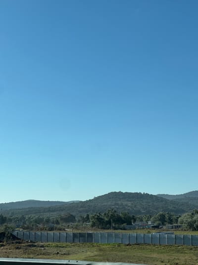The image displays an expansive, clear blue sky occupying approximately the upper two-thirds of the frame. The lower third features a landscape, observed from a low vantage point, possibly from within a vehicle, indicated by a metallic silver rail component visible at the bottom edge.
In the midground, a uniform barrier composed of numerous rectangular, light-gray concrete panels runs horizontally across the scene. Each panel is vertically oriented and evenly spaced, forming a continuous line. Behind this concrete barrier, a landscape unfolds with scattered green trees and some rudimentary, low-rise structures with light-colored walls, indicative of ongoing construction or established simple buildings. Utility poles or similar vertical structures are visible amidst the trees and buildings.
Further in the background, a series of rolling hills, densely covered with dark green vegetation, recede towards the horizon under the bright sky. The immediate foreground consists of a narrow strip of dry, patchy green and brown vegetation. No individuals, specific actions, or interactions are discernible within the scene. The overall environment suggests a rural-urban interface. This scene is located in the vicinity of Amozoc de Mota, Mexico.