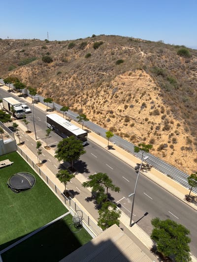 The image depicts a sunny day in Sderot, Israel, showing a road bordering a dry, scrub-covered hillside. A white bus is traveling on the road, with a white truck and a white car parked on the left side of the road. The roadside features several young trees and a sidewalk. To the left of the road, a fenced area contains a green lawn with a trampoline and a bench. In the foreground, a section of a building's roof is visible, casting a shadow. The hillside is rocky and dry, with sparse vegetation and a fence along its base. The sky is clear and blue.