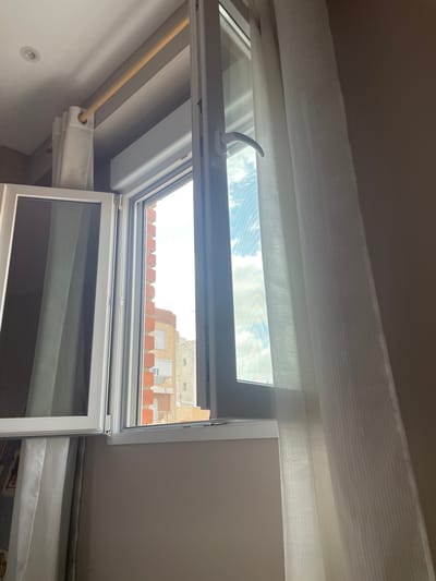 The image shows an interior view looking upwards towards an open skylight window. The window is partially open, allowing a view of a blue sky with white clouds. Outside the window, the upper floors of buildings with terracotta tile roofs are visible. The scene appears to be set in Sagunto, Spain, as indicated by the architectural style and rooftops.
Inside, the skylight window has a white frame and a white handle for opening and closing. Below the skylight, there is a white frame with what appears to be a screen or a dark-tinted glass. There are also white blinds or curtains partially drawn. A wooden rod is visible, extending from the left side of the frame and threaded through a loop in the white material, suggesting it is used to operate the blinds or curtains. The walls and ceiling are painted in neutral tones.
The lighting suggests it is daytime, possibly late morning or afternoon, with bright sunlight filtering through the window. The overall impression is that of a residential interior, perhaps an attic room or a room with a sloped ceiling, featuring a functional and modern skylight. There is no visible text in the image.