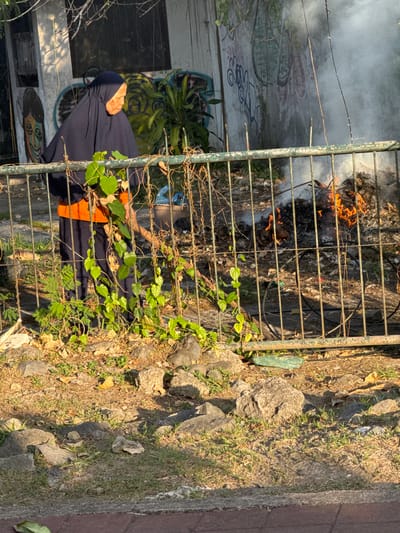 A woman wearing a dark blue hijab and an orange shirt stands behind a metal fence, tending to a small bonfire. The fire is producing a plume of white smoke that rises into the air and obscures a graffiti-covered wall in the background. The area around the fire appears to be littered with dried leaves and debris. The ground is covered with grass, rocks, and a paved walkway in the foreground. The scene is lit by the warm light of what appears to be a sunset or sunrise. The location is the city of Denpasar Barat, Indonesia.