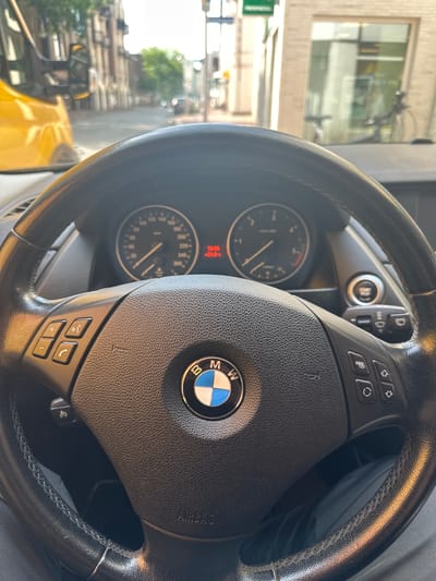 This is a first-person perspective shot from inside a car, looking forward through the windshield. The foreground is dominated by a black leather BMW steering wheel with visible controls for volume, phone, and cruise control. The BMW logo is prominently displayed in the center of the steering wheel. Beyond the steering wheel, the car's dashboard displays a speedometer and a digital information screen showing "10:05" and "-24.0°".
The exterior view through the windshield shows a street scene in a European city. A bright yellow vehicle, possibly a van, is partially visible on the left. The street is lined with buildings, suggesting an urban environment. Trees with green foliage are present, indicating it is likely daytime and a season with leaves. The buildings appear to be a mix of traditional architecture with brick facades and some modern elements. The overall impression is that of a sunny day in a city.
Based on the license plate visible on the yellow vehicle on the left (partially obscured but with letters that could potentially be Gütersloh related, such as 'GT' or similar), and the architectural style of the buildings, the location is likely Gütersloh, Germany. However, without a clearer view of the license plate or identifying street signs, this is an educated inference.