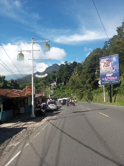 The image depicts a daytime roadside scene in the city of Ka Thaung (Upper), Myanmar. A two-lane asphalt road, marked with a solid white line on the left edge and a dashed yellow line further right, extends diagonally from the lower left foreground towards the upper center-right. Long shadows from overhead wires stretch across the road surface.
On the immediate left, a white decorative street lamp pole features two ornate, cage-like lanterns and a central circular sign. This sign displays Arabic script above the Latin text "AL ALIIM." Behind the pole, a series of rustic roadside stalls with corrugated metal roofs, some rusted, and blue/brown painted walls are visible along a dirt shoulder. Multiple motorcycles are parked near these stalls, with one motorcycle featuring a purple covering. At least two individuals are present near the parked motorcycles and stalls. Further down this side, a white SUV is also parked.
On the right side of the road, a large blue billboard stands prominently. It advertises "Old Italian Village" and "ROOF PARK," indicates a distance of "4.7 Km," and lists "Puncak, Bogor, Cianjur" as geographical references. Beyond the billboard, a steep, densely forested mountain slope rises, with a distant white multi-story building partially visible amidst the trees.
The road itself carries traffic, consisting of a line of several motorcycles and at least one car, all moving away from the viewer. The background is dominated by lush green mountains under a bright blue sky dotted with scattered white clouds.