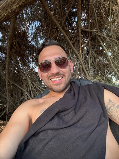 A man wearing aviator sunglasses and a black patterned blanket smiles at the camera. He has a light beard and dark hair. The background includes a tree with many bare branches reaching upwards. Tattoos are visible on his arm and shoulder. The image is taken from a low angle, looking upwards towards the man and tree.