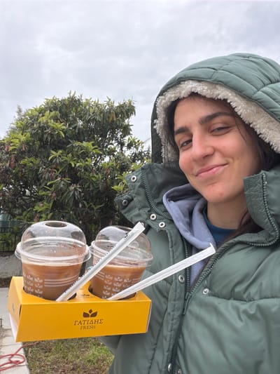 A person wearing a green hooded jacket with a white fleece lining and a blue sweatshirt underneath is holding a yellow cardboard carrier containing two clear plastic cups of a brown beverage with white straws. The carrier has the word "ΓΑΤΙΔΗΣ" and "FRESH" printed vertically on its side. The person is smiling and looking down at the drinks. Behind the person, there is a tree with green leaves, a cloudy sky, and some ground with sparse vegetation. The location is inferred to be Megas Alexandros, Greece, based on the presence of the Greek text on the carrier. The image is slightly tilted.