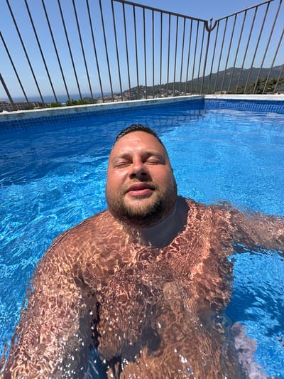 A man with closed eyes, a beard, and short dark hair is submerged in a bright blue swimming pool on a sunny day. Water covers most of his chest and shoulders, refracting light and creating rippling patterns on his skin. In the background, a metal fence with vertical bars overlooks a coastal town with buildings clustered on a hillside, leading down to the blue sea. The sky is clear and blue. This scene appears to be from a rooftop or elevated pool in Sant Feliu de Guíxols, Spain, given the distinctive coastal landscape and architecture.