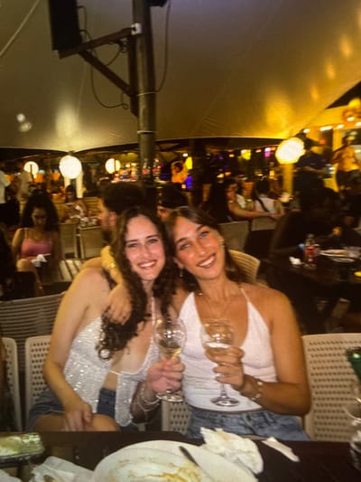 This image displays two young adult females posing for a photograph in a crowded outdoor or semi-outdoor restaurant or bar located in Cairu, Brazil, at night. The female on the left, possessing long, dark, curly hair, is wearing a light-colored, possibly sequined, halter-neck top and denim shorts. Her right arm is around the shoulder of the female on the right. She holds a wine glass containing a clear liquid in her left hand. The female on the right, with medium-length brown hair, wears a light-colored halter-neck top and light-wash denim shorts, holding a wine glass with a clear liquid in her right hand. Both individuals are smiling towards the camera. On the dark table in the immediate foreground, a plate with food residue and crumpled white napkins is visible. The background reveals numerous blurred patrons seated at various tables, indicating a busy environment. The area is illuminated by warm artificial lighting, including spherical white lamps and multi-colored lights. Overhead, a large, light-colored canopy or tent structure is visible, supported by a central pole. A male figure's back and shoulder are discernible between the two foreground subjects. Further into the background, a female in a pink top is seated, and a person in a yellow shirt is partially visible in the far right, possibly on an elevated platform.