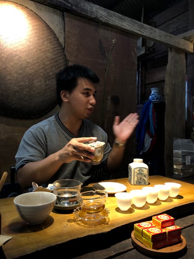 A man is sitting at a wooden table in what appears to be a tea house in B'Lao, Vietnam. He is wearing a grey t-shirt and holding a tea cup in his right hand, gesturing with his left hand. On the table in front of him are several tea cups, a tea pot, and some boxes of what appear to be tea. The background is rustic, with wooden walls and a woven wall hanging. There is a fern growing in a pot in the background. The lighting is dim, with a spotlight on the woven wall hanging.