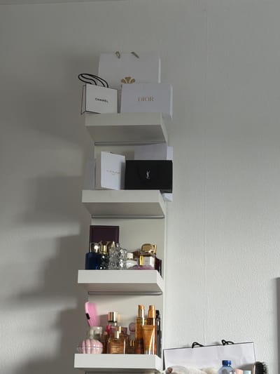 The image displays an organized collection of luxury brand items and beauty products arranged on white, wall-mounted shelves in an indoor setting, likely in Groningen, Netherlands. The wall is light grey or white with a textured finish.
The shelving unit is modular, comprising several white rectangular compartments. On the leftmost section, a white Dior shopping bag with gold details and a white box with "DIOR" text are visible. Below them, a small white Chanel shopping bag with "CHANEL" text is hanging.
The central sections of the shelving hold a black box featuring the "YSL" logo (Yves Saint Laurent) and a white box labeled "GUERLAIN PARIS".
The rightmost sections are filled with an assortment of perfume bottles and beauty product containers, many with golden accents, suggesting a collection of fragrances or cosmetics. A dark red wallet or small clutch is also present. Further to the right, a pink hairbrush and a light pink, shell-shaped item are visible. Partially seen on the far right are a white shopping bag with black handles, a blue hairbrush, a small plastic water bottle, and a white towel or fabric.
No individuals are present, and no activity is taking place other than the static display of items. The lighting appears to be artificial, casting soft shadows beneath the shelves on the wall. Visible text includes "DIOR", "CHANEL", the "YSL" logo, "GUERLAIN PARIS", and "NUXE" on a product tube. The arrangement suggests a personal space for storing and displaying high-end items.