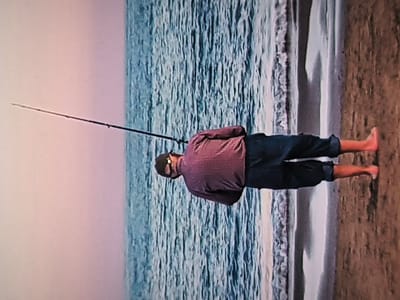 The image, when correctly oriented, depicts an adult male standing on a sandy beach at the water's edge, facing the ocean. He is viewed from behind and is holding a fishing rod that extends out over the water, indicating he is engaged in fishing.
The man is wearing a reddish-purple long-sleeved shirt, dark blue trousers or shorts rolled up to his calves, a dark baseball cap, and sunglasses. He is barefoot, standing where the gentle waves meet the shore. The ocean stretches into the distance, appearing textured with ripples and reflections. Small, foamy waves can be seen breaking softly onto the sand. The sky above presents a soft gradient of light pink and purple hues, suggesting either dawn or dusk. The scene conveys a sense of tranquility. No text is visible in the image.