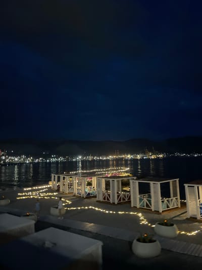 The image shows an outdoor seating area at night, possibly a restaurant or lounge, situated by a body of water. White gazebos with slatted walls and open fronts are arranged on a raised platform. Each gazebo appears to have seating inside. The area is illuminated by strings of warm-toned lights that are draped along the platform and around the gazebos. Two people are visible on the platform, one standing near a gazebo and the other walking away from the camera. In the foreground, there are low, rectangular tables. The background features a dark body of water with reflections of distant lights from a city or town lining the shore. Mountains are visible in the far distance, silhouetted against the dark night sky. The overall atmosphere is serene and ambient, suggesting a place for relaxation or dining.