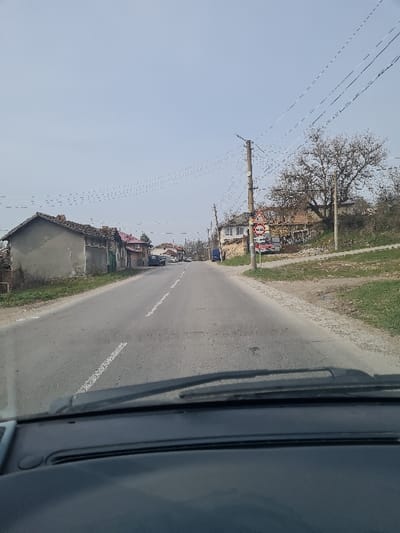 The image depicts a two-lane asphalt road in Blagoevgrad, Bulgaria, viewed from inside a vehicle through its windshield on a clear day. The road, exhibiting surface wear and a faded white dashed center line, extends into the distance. On the left side, a series of residential structures, including a prominent single-story building with a light grey, unrendered facade and a tiled roof, line the road. Multiple overhead power lines are visible, strung between utility poles on both sides of the thoroughfare. On the right side, a grassy verge transitions into higher ground where a utility pole stands, displaying a red-bordered circular road sign with a white background and black horizontal bar, indicating "No Entry for All Vehicles." Behind this pole, a building undergoing construction or renovation is observable, alongside a large deciduous tree with bare branches. The dark dashboard and two windshield wipers of the observer's vehicle occupy the lower portion of the frame. No individuals or active movements are discernible.