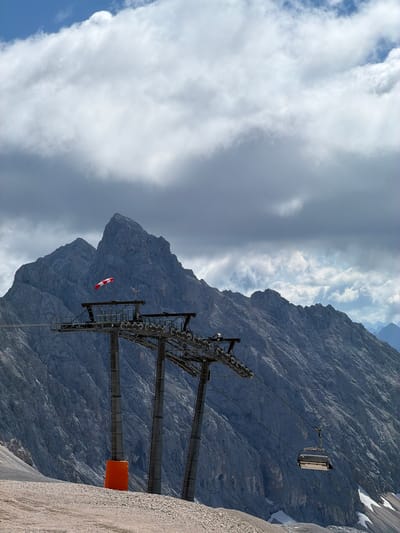 This is a photograph of a mountain landscape with a cable car system in the foreground. The scene is dominated by a large, rugged mountain face with visible rock formations and patches of snow. The sky is filled with a mix of white and gray clouds, suggesting a partly cloudy day.
The cable car system includes large support towers, wires, and a cable car suspended on one of the lines. The cable car itself appears to be a modern design, enclosed and metallic. A windsock, red and white striped, is visible hanging from one of the support structures, indicating wind conditions. The ground in the lower right of the frame appears to be a gravelly, mountainous terrain.
The time of day is likely daytime, given the lighting on the mountains and clouds. The weather appears to be dynamic, with both clear sky and heavy cloud cover present. The mountainous terrain and the cable car suggest this is a location in the Alps, specifically Wildermieming, Austria, likely a ski resort or a popular mountain destination. There are no people visible in the image, and the overall mood is one of natural grandeur and possibly outdoor adventure.