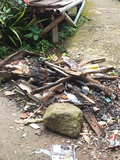 A chaotic pile of debris rests on the ground near a simple bamboo bench. The refuse includes burnt wood, charred fragments, and pieces of corrugated metal. A large, moss-covered rock sits among the mess. Litter, such as wrappers and a discarded carton, adds to the untidiness. Greenery and plants grow near the bench, contrasting with the surrounding disarray.