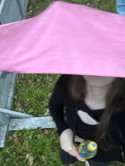 The image shows a person holding a yellow and blue bottle, partially obscured by a pink tarp. The person is wearing a black top with a cut-out detail on the chest. They are outdoors, with grass and a wooden fence visible in the background. The overall setting suggests a casual outdoor gathering or event. No specific details about the city of Nevyansk, Russia, can be inferred from the image.