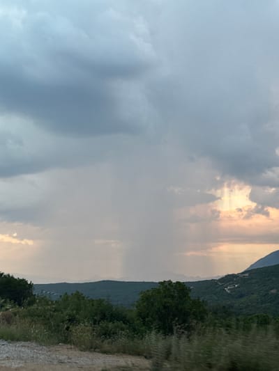 The image depicts a mountainous landscape with a dramatic sky. In the foreground, there is a gravelly path and dense green foliage. The middle ground is dominated by rolling hills covered in lush vegetation, with a winding road and scattered buildings visible on the right slope. The background shows a hazy mountain range stretching across the horizon. The sky is filled with heavy, gray clouds, with shafts of light breaking through on the right, suggesting rain is falling in the distance. The overall impression is one of natural beauty and atmospheric conditions typical of Višnjevo, Montenegro.